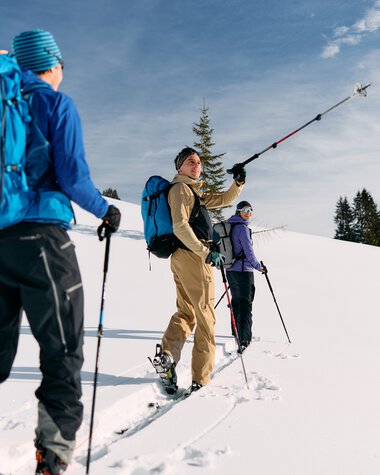 Drei Skitourengeher mit Rucksäcken in Schneelandschaft, einer macht eine selfie-artige Aufnahme während einer Wintertour im Kleinwalsertal. | © Kleinwalsertal Tourismus eGen • Fotograf: Oliver Farys