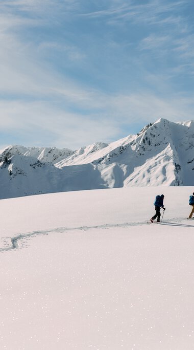 Drei Skitourengeher unterwegs in verschneiter Berglandschaft im Kleinwalsertal, umgeben von hohen, schneebedeckten Gipfeln bei schönem Wetter. | © Kleinwalsertal Tourismus eGen • Fotograf: Oliver Farys