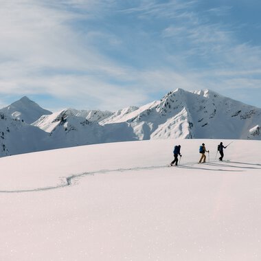 Drei Skitourengeher unterwegs in verschneiter Berglandschaft im Kleinwalsertal, umgeben von hohen, schneebedeckten Gipfeln bei schönem Wetter. | © Kleinwalsertal Tourismus eGen • Fotograf: Oliver Farys