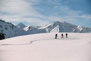 Drei Skitourengeher unterwegs in verschneiter Berglandschaft im Kleinwalsertal, umgeben von hohen, schneebedeckten Gipfeln bei schönem Wetter. | © Kleinwalsertal Tourismus eGen • Fotograf: Oliver Farys