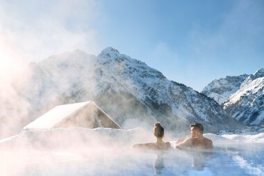 Zwei Personen entspannen sich in einem Hot Tub inmitten von verschneiten Bergen im Kleinwalsertal. | © Kleinwalsertal Tourismus eGen • Fotograf: Oliver Farys