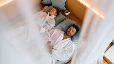 © Kleinwalsertal Tourismus eGen • Fotograf: Oliver Farys Ein Mann und eine Frau liegen im Wellness-Spa-Bereich auf Liegen, eingekuschelt in Bademäntel, mit Kissen und einer Tasse. Es wirkt entspannt und ruhig. | © Kleinwalsertal Tourismus eGen • Fotograf: Oliver Farys