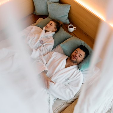 Ein Mann und eine Frau liegen im Wellness-Spa-Bereich auf Liegen, eingekuschelt in Bademäntel, mit Kissen und einer Tasse. Es wirkt entspannt und ruhig. | © Kleinwalsertal Tourismus eGen • Fotograf: Oliver Farys