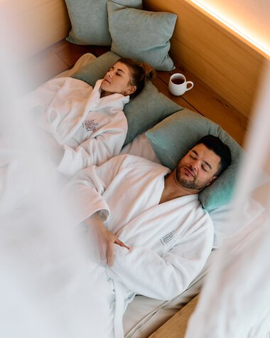 Ein Mann und eine Frau liegen im Wellness-Spa-Bereich auf Liegen, eingekuschelt in Bademäntel, mit Kissen und einer Tasse. Es wirkt entspannt und ruhig. | © Kleinwalsertal Tourismus eGen • Fotograf: Oliver Farys