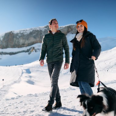 Zwei Personen wandern bei winterlichem Schneewetter im Kleinwalsertal, begleitet von einem Hund, mit Bergen im Hintergrund. | © Kleinwalsertal Tourismus eGen • Fotograf: Oliver Farys