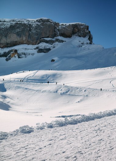 Zwei Personen mit Hund wandern im verschneiten Gebirge im Kleinwalsertal bei schönem Winterwetter. | © Kleinwalsertal Tourismus eGen • Fotograf: Oliver Farys