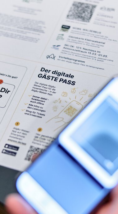 Person hält einen digitalen Gästepass und liest ein gedrucktes Dokument bei einem Tisch, eine Person fotografiert es mit einem Smartphone. | © Kleinwalsertal Tourismus eGen • Fotograf: Oliver Farys