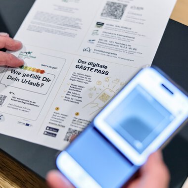 Person hält einen digitalen Gästepass und liest ein gedrucktes Dokument bei einem Tisch, eine Person fotografiert es mit einem Smartphone. | © Kleinwalsertal Tourismus eGen • Fotograf: Oliver Farys