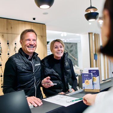 Zwei lachende Menschen beim Empfang am Winter 2025-Event im Kleinwalsertal, hinter dem Tresen im Tourismusbüro. | © Kleinwalsertal Tourismus eGen • Fotograf: Oliver Farys