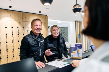Zwei lachende Menschen beim Empfang am Winter 2025-Event im Kleinwalsertal, hinter dem Tresen im Tourismusbüro. | © Kleinwalsertal Tourismus eGen • Fotograf: Oliver Farys