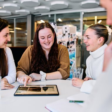 Four young women sitting around a table, smiling and looking at a tablet in a modern office. | © Kleinwalsertal Tourismus eGen • Photographer: Oliver Farys