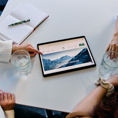 Vier Personen sitzen um einen Tisch und schauen auf ein Tablet mit einer Berglandschaft auf dem Bildschirm, im Büro während eines Meetings. | © Kleinwalsertal Tourismus eGen • Fotograf: Oliver Farys