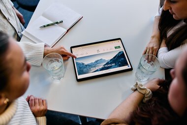 Vier Personen sitzen um einen Tisch und schauen auf ein Tablet mit einer Berglandschaft auf dem Bildschirm, im Büro während eines Meetings. | © Kleinwalsertal Tourismus eGen • Fotograf: Oliver Farys
