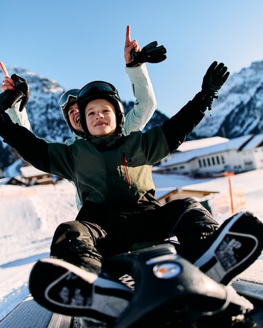 Kinder beim Rodeln im Kleinwalsertal, fröhlich lachend, mit Bergen im Hintergrund, Winterlandschaft. | © Kleinwalsertal Tourismus eGen • Fotograf: Oliver Farys