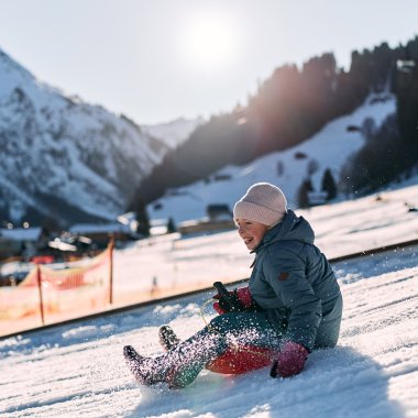 Ein Kind sitzt lachend in schmelzendem Schnee bei Sonnenlicht, umgeben von Bergen, während es rodeln geht. | © Kleinwalsertal Tourismus eGen • Fotograf: Oliver Farys