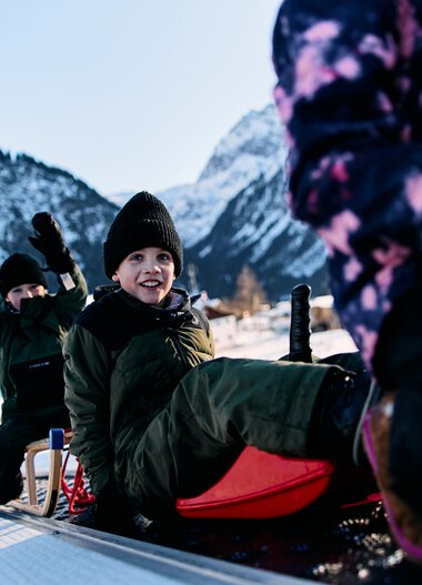 Kinder beim Rodeln im Schnee in den Bergen, lachend und spielend, Winterurlaub im Kleinwalsertal. | © Kleinwalsertal Tourismus eGen • Fotograf: Oliver Farys