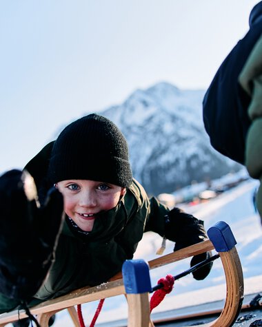 Kind beim Rodeln im Kleinwalsertal, lachend und in Wintersportkleidung, mit Bergen im Hintergrund. | © Kleinwalsertal Tourismus eGen • Fotograf: Oliver Farys