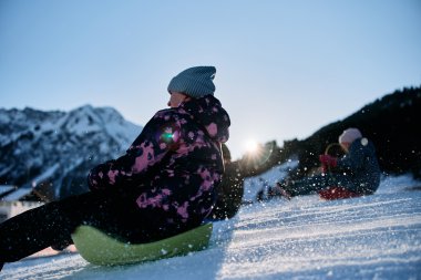 Kinder beim Rodeln in den Bergen im Kleinwalsertal, sonniger Wintertag mit Schneelandschaft, im Hintergrund Gipfel. | © Kleinwalsertal Tourismus eGen • Fotograf: Oliver Farys