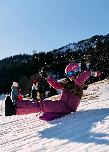 Kinder beim Rodeln im Winter in Kleinwalsertal, lachend und spielend auf einem verschneiten Hang, umgeben von Bergen. | © Kleinwalsertal Tourismus eGen • Fotograf: Oliver Farys