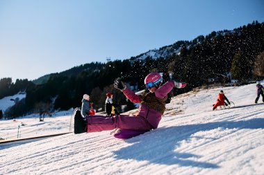 Kinder beim Rodeln im Winter in Kleinwalsertal, lachend und spielend auf einem verschneiten Hang, umgeben von Bergen. | © Kleinwalsertal Tourismus eGen • Fotograf: Oliver Farys