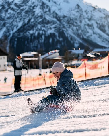 Ein Kind sitzt im Schnee beim Rodeln im Kleinwalsertal, umgeben von Bergen und Wintersportlern bei sonnigem Wetter. | © Kleinwalsertal Tourismus eGen • Fotograf: Oliver Farys