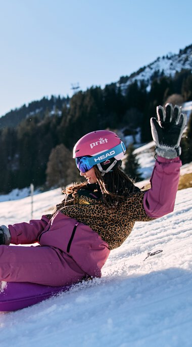 Ein Mädchen rutscht lachend und lachend auf einem Schneehang im Kleinwalsertal, umgeben von Bergen und anderen Kindern beim Rodeln, bei sonnigem Winterwetter. | © Kleinwalsertal Tourismus eGen • Fotograf: Oliver Farys