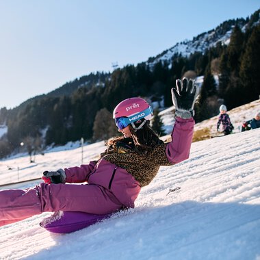 Ein Mädchen rutscht lachend und lachend auf einem Schneehang im Kleinwalsertal, umgeben von Bergen und anderen Kindern beim Rodeln, bei sonnigem Winterwetter. | © Kleinwalsertal Tourismus eGen • Fotograf: Oliver Farys