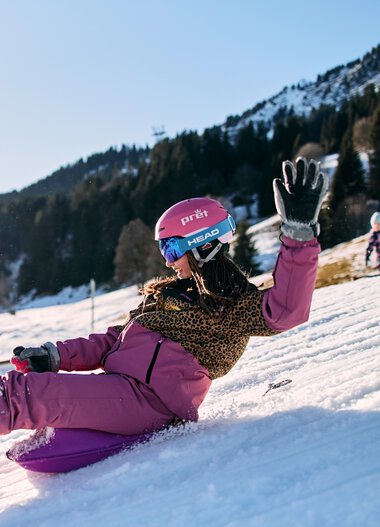 Ein Mädchen rutscht lachend und lachend auf einem Schneehang im Kleinwalsertal, umgeben von Bergen und anderen Kindern beim Rodeln, bei sonnigem Winterwetter. | © Kleinwalsertal Tourismus eGen • Fotograf: Oliver Farys