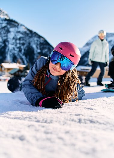 Mädchen liegt auf Schnee im Skigebiet, trägt pinken Helm und Skibrille, im Hintergrund Berge und weitere Skifahrer. | © Kleinwalsertal Tourismus eGen • Fotograf: Oliver Farys