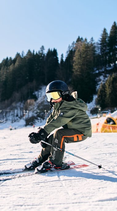 Ein Kind beim Skifahren im Kleinwalsertal im Winter 2025, trägt einen Helm und eine Skiausrüstung, umgeben von Schneelandschaft und Bäumen. | ©  Kleinwalsertal Tourismus eGen • Fotograf: Oliver Farys