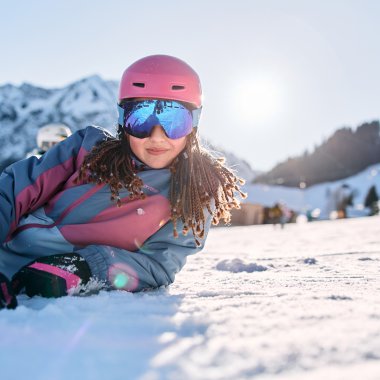 Mädchen beim Skifahren in den Bergen, trägt pinken Helm und Skibrille, liegt im Schnee, im Hintergrund verschneite Berge. | © Kleinwalsertal Tourismus eGen • Fotograf: Oliver Farys
