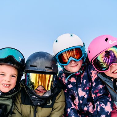 Vier Kinder beim Skifahren in Winterkleidung und Helmen, lachen und posieren im Schnee. | © Kleinwalsertal Tourismus eGen • Fotograf: Oliver Farys