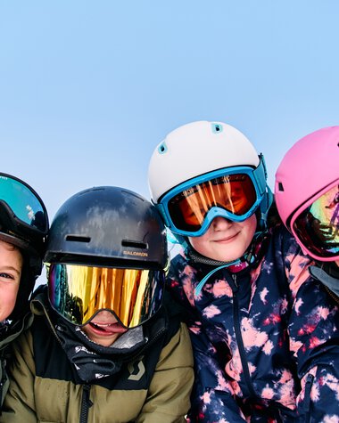 Vier Kinder beim Skifahren in Winterkleidung und Helmen, lachen und posieren im Schnee. | © Kleinwalsertal Tourismus eGen • Fotograf: Oliver Farys