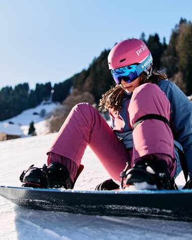 Junge Skifahrerin in pinker Skijacke und Helm sitzt auf dem Snowboard im Schnee, umgeben von Winterlandschaft mit schneebedecktem Berg und Bäumen. | © Kleinwalsertal Tourismus eGen • Fotograf: Oliver Farys