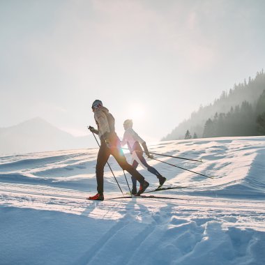 Zwei Skifahrer beim klassischen Langlauf auf der Schwarzwasserloipe im Kleinwalsertal bei Sonnenaufgang, umgeben von verschneiter Landschaft und Bergen. | © Kleinwalsertal Tourismus eGen • Fotograf: Oliver Farys