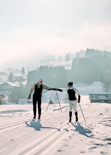 Zwei Langläufer auf der Winter-Loipe im Kleinwalsertal, im Hintergrund schneebedeckte Häuser und Berge, bewölkter Himmel, Winterlandschaft. | © Kleinwalsertal Tourismus eGen • Fotograf: Oliver Farys