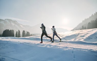 Zwei Skifahrer ziehen im frühen Morgenlicht durch die verschneite Schwarzwasserloipe im Kleinwalsertal, umgeben von Bergen und Tannen | © Kleinwalsertal Tourismus eGen • Fotograf: Oliver Farys