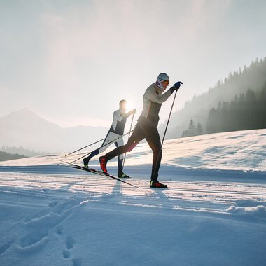 Zwei Skilangläufer in winterlicher Landschaft, Sonne im Hintergrund, auf einer Schneepiste im Kleinwalsertal. | © Kleinwalsertal Tourismus eGen • Fotograf: Oliver Farys