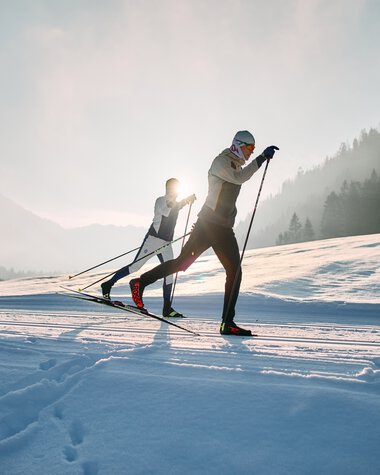 Zwei Skilangläufer in winterlicher Landschaft, Sonne im Hintergrund, auf einer Schneepiste im Kleinwalsertal. | © Kleinwalsertal Tourismus eGen • Fotograf: Oliver Farys