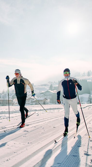 Zwei Skifahrer laufen auf einer verschneiten Langlaufloipe im Kleinwalsertal mit Bergen im Hintergrund. | © Kleinwalsertal Tourismus eGen • Fotograf: Oliver Farys