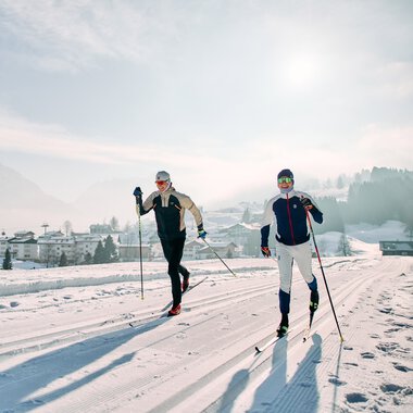Zwei Skifahrer laufen auf einer verschneiten Langlaufloipe im Kleinwalsertal mit Bergen im Hintergrund. | © Kleinwalsertal Tourismus eGen • Fotograf: Oliver Farys