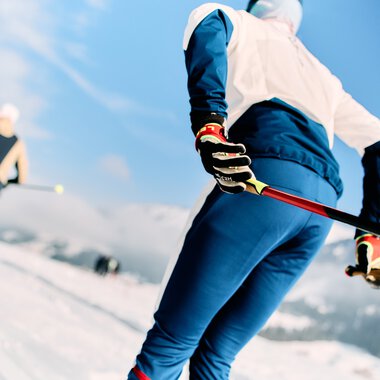 Cross-country skiers in a snowy landscape with mountains and blue sky in the background | © Kleinwalsertal Tourismus eGen • Photographer: Oliver Farys