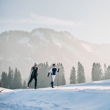 Zwei Skilangläufer auf der Schwarzwasserloipe in den österreichischen Alpen, umgeben von verschneiten Bergen und Tannen, bei winterlicher Kulisse. | © Kleinwalsertal Tourismus eGen • Fotograf: Oliver Farys