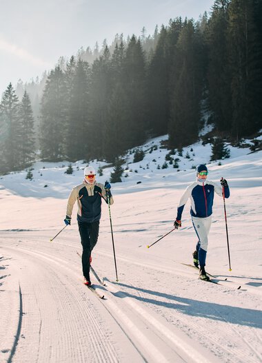 Two cross-country skiers skiing on the Schwarzwasserloipe in Kleinwalsertal, surrounded by snow-covered trees and mountains in a winter landscape. | © Kleinwalsertal Tourismus eGen • Photographer: Oliver Farys