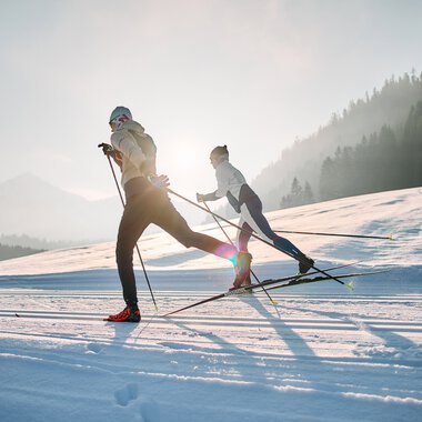 Two cross-country skiers on the Schwarzwasserloipe in Kleinwalsertal, surrounded by mountains and forest at sunrise. | © Kleinwalsertal Tourismus eGen • Photographer: Oliver Farys