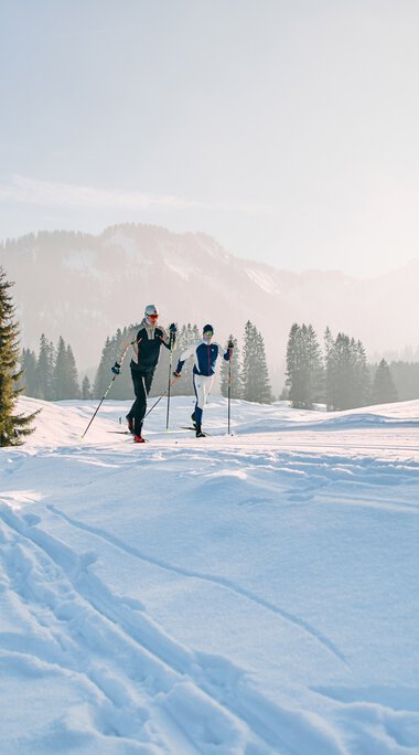 Zwei Menschen beim Langlaufen in winterlicher Landschaft mit Schnee, Holzhaus und Tannenbäumen im Hintergrund. | © Kleinwalsertal Tourismus eGen • Fotograf: Oliver Farys