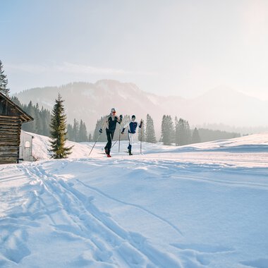 Zwei Menschen beim Langlaufen in winterlicher Landschaft mit Schnee, Holzhaus und Tannenbäumen im Hintergrund. | © Kleinwalsertal Tourismus eGen • Fotograf: Oliver Farys