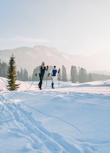 Zwei Menschen beim Langlaufen in winterlicher Landschaft mit Schnee, Holzhaus und Tannenbäumen im Hintergrund. | © Kleinwalsertal Tourismus eGen • Fotograf: Oliver Farys