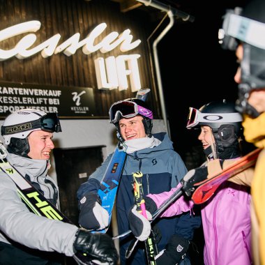 Vier Skifahrer in Winterkleidung und Helm, lachend und im Gespräch, bei Nacht in einem Skigebiet, Hintergrunde mit Leuchtschrift 'Kaiser Lift' und einem Ankündigungsschild. | © Kleinwalsertal Tourismus eGen • Fotograf: Oliver Farys