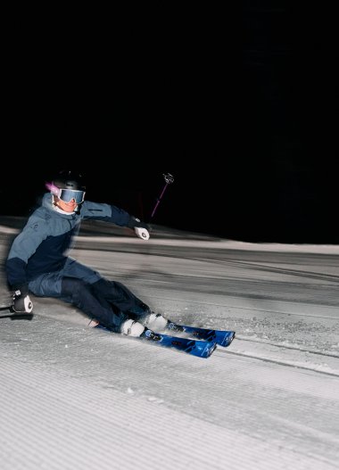 Skifahrer bei Nacht im Kleinwalsertal, der eine enge Kurve im Schnee fährt, mit dunklem Himmel im Hintergrund. | © Kleinwalsertal Tourismus eGen • Fotograf: Oliver Farys